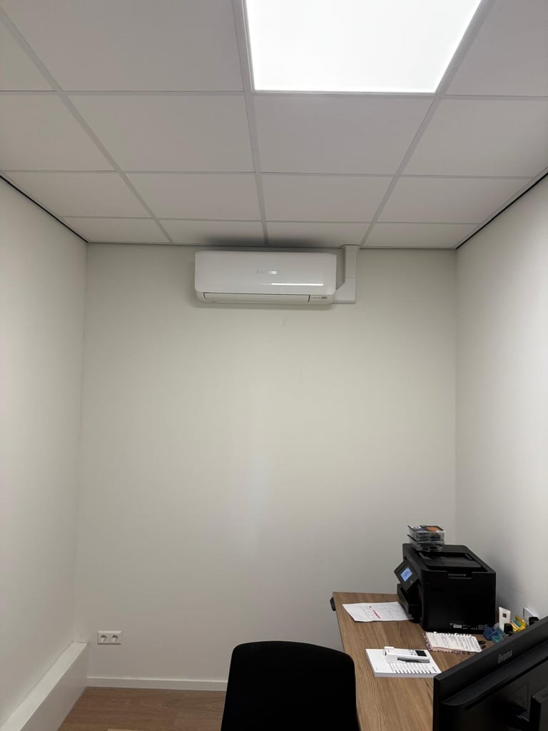 Small office room with white walls, drop ceiling with recessed lighting, wall-mounted air conditioning unit, wooden desk with office equipment, and black office chair