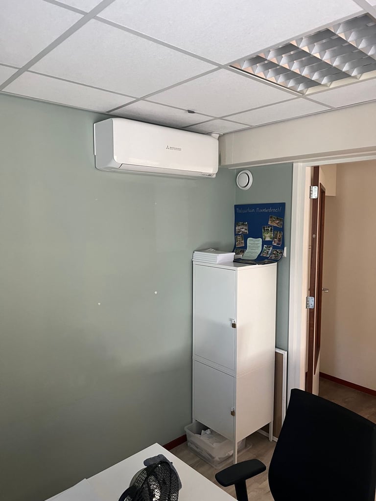 Office room with mint green wall, white air conditioning unit mounted above, white storage cabinet, and ceiling with fluorescent lights