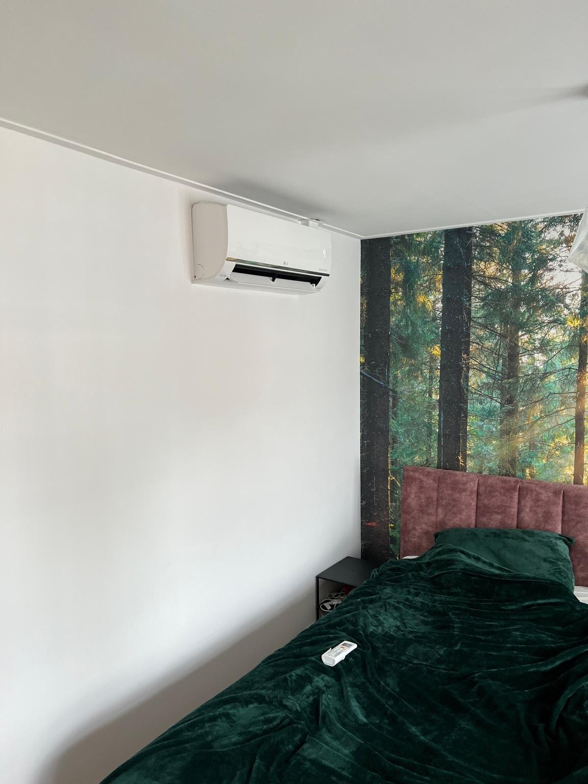 Modern bedroom with green velvet bedding, mauve headboard, white walls, air conditioning unit, and forest wallpaper accent