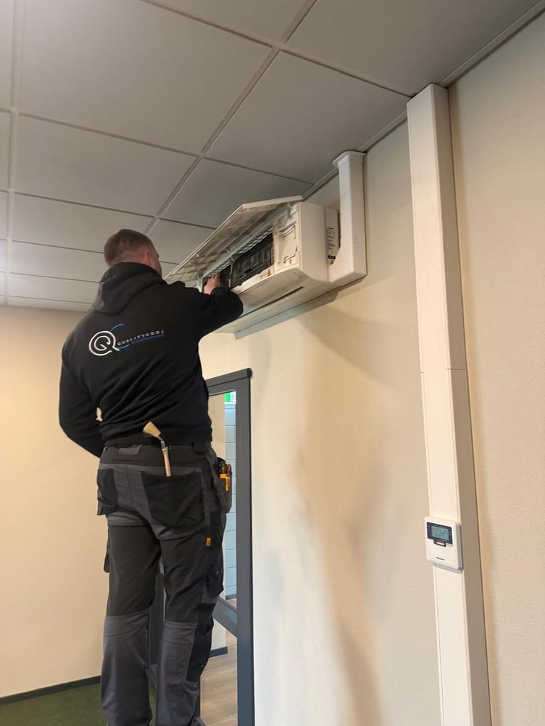 Technician in black jacket reaching up to service an air conditioning unit mounted on a wall.