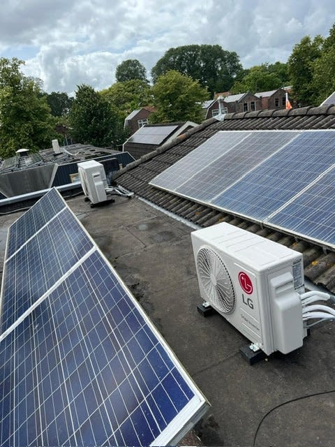 Solar panels installed on a residential rooftop with an air conditioning unit, surrounded by brick buildings and green trees under a cloudy sky