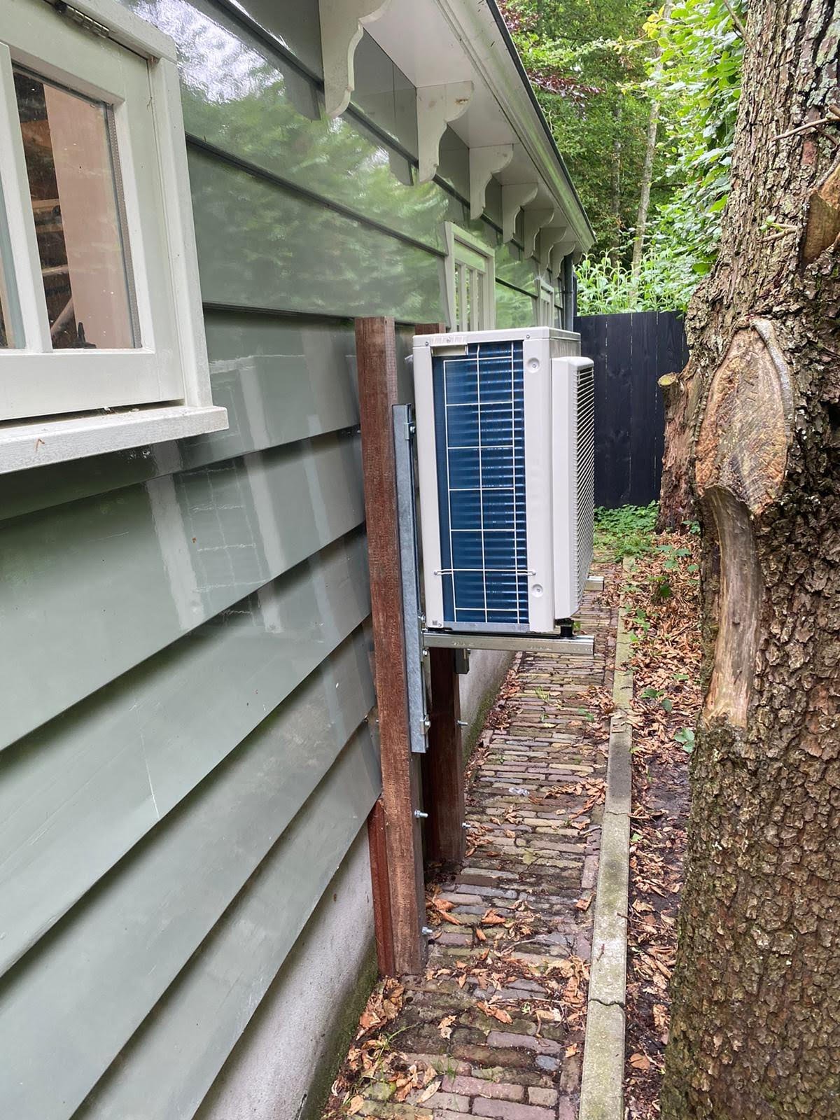 Outdoor air conditioning unit mounted on wooden stand beside house with green siding and brick pathway