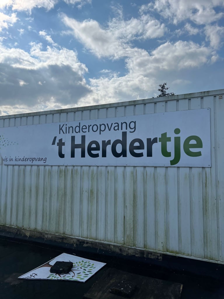 White metal sign for Kinderopvang t Herdertje daycare center with blue sky and white clouds behind it