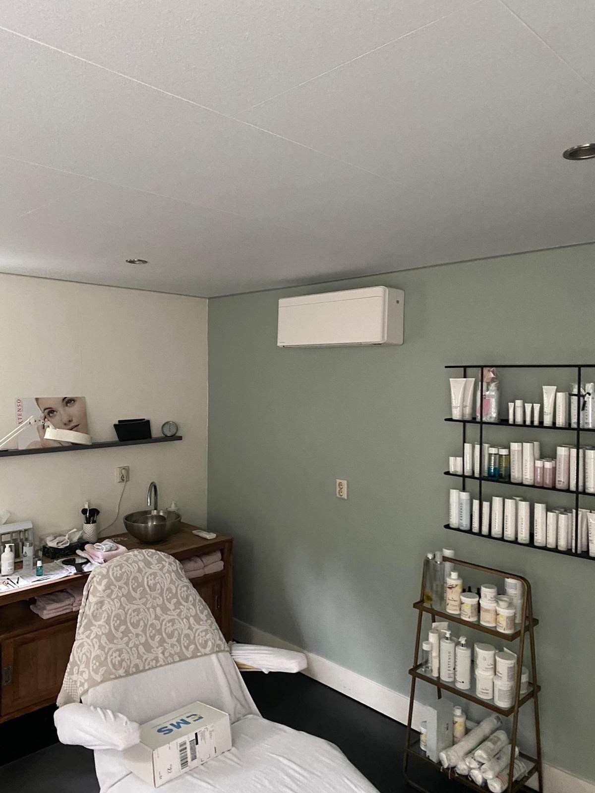 A beauty treatment room with white bed, wooden desk, wall-mounted shelving with products, and pale green walls