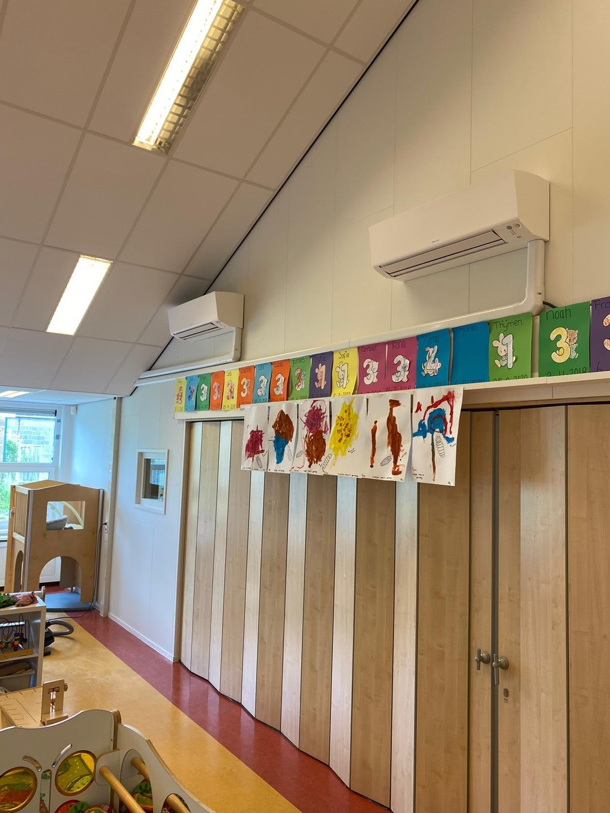 Classroom interior with colorful alphabet posters on beige walls above wooden storage cubbies and doors, fluorescent ceiling lights, and play area visible on left