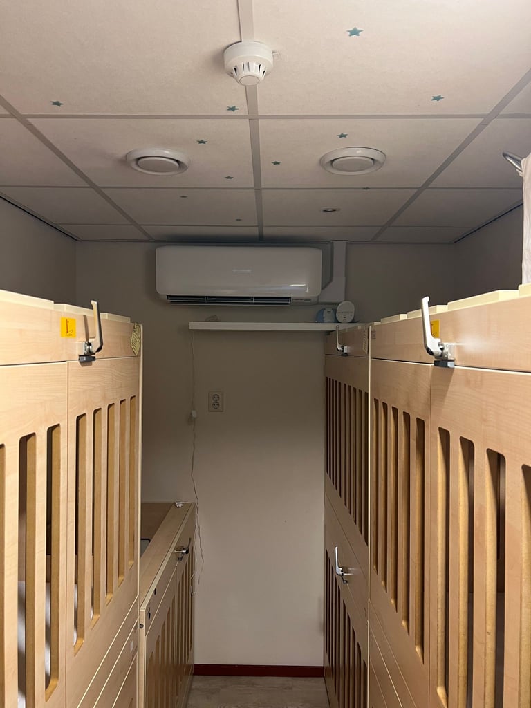 Institutional nursery room with two wooden cribs along side walls, suspended ceiling lights, air conditioning unit, and recessed lighting