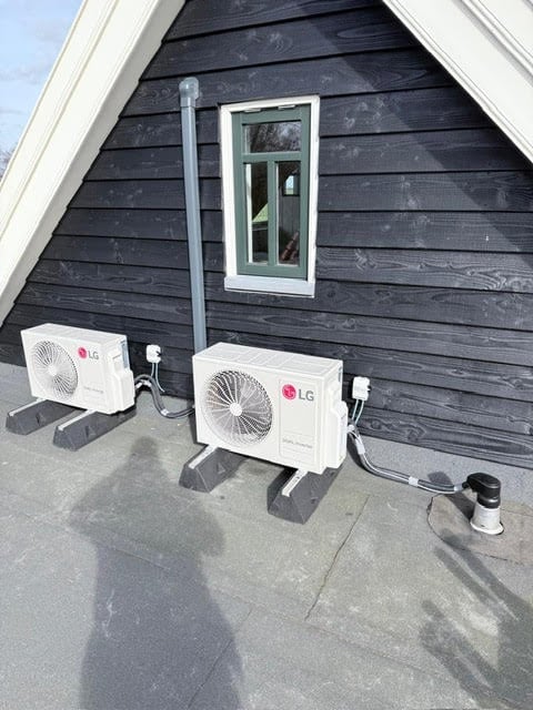 Three LG heat pump units mounted on concrete pad against dark wooden house wall with white trim window