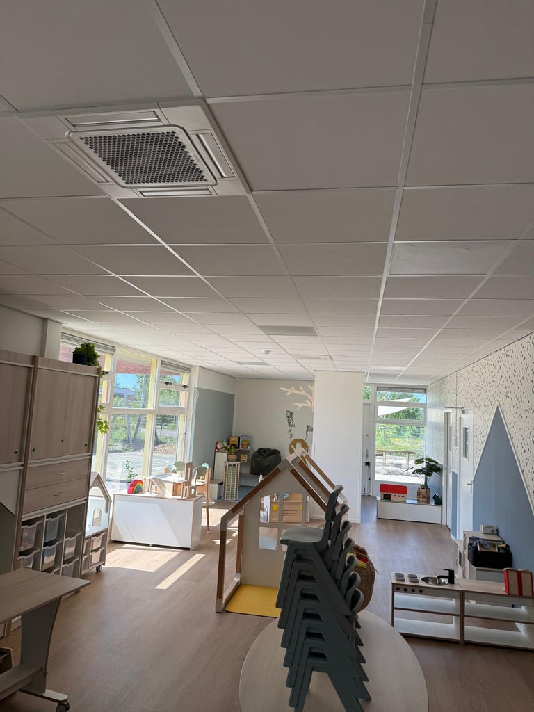 Interior of a daycare or classroom with drop ceiling, wooden playhouse structure, windows with natural light, and various furniture and toys