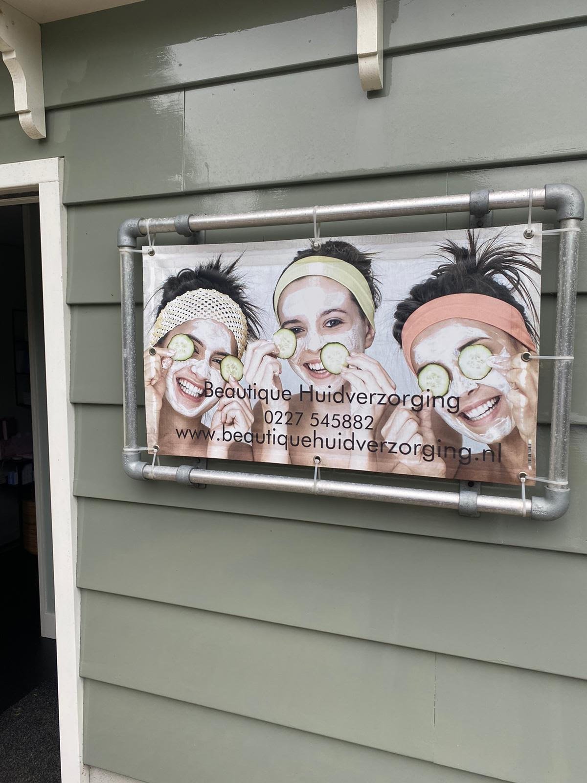 Business sign for Beautique Huidverzorging mounted on green siding, showing three women with spa treatments and contact information