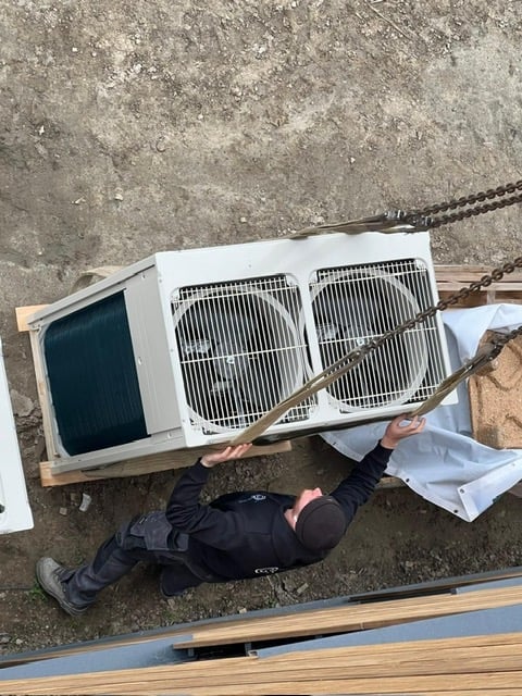 Technician lying on concrete ground working on white air conditioning condenser unit with exposed coils and metal fins
