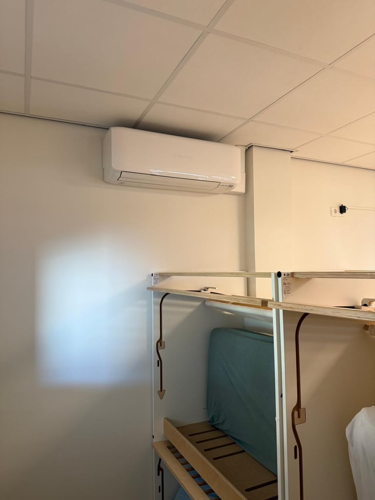 Dorm room interior with wall-mounted air conditioning unit above a metal bunk bed frame with teal mattress